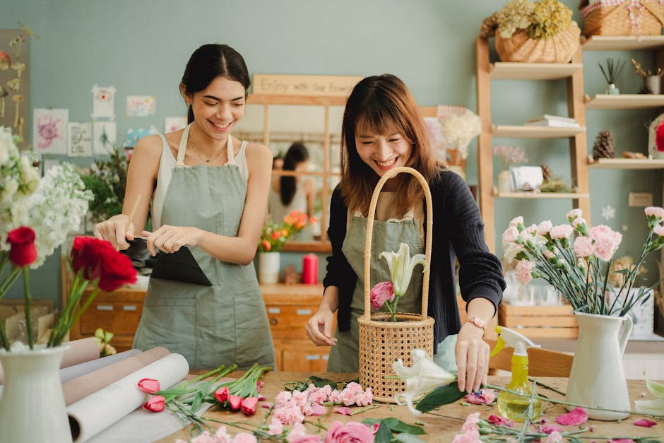 Two women working in a florist shop, assembling a floral gift arrangement. The woman on the left is wearing a light green apron and holding a black wrapping cloth, smiling as she looks down at her wor