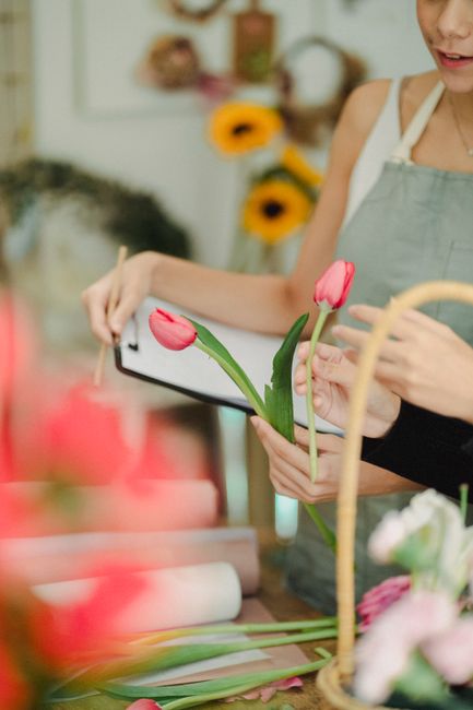 A florist at Walworth Florist is holding a vibrant pink tulip with green stems and leaves, examining it closely while preparing a floral arrangement. The florist’s hands are visible, and she is wear