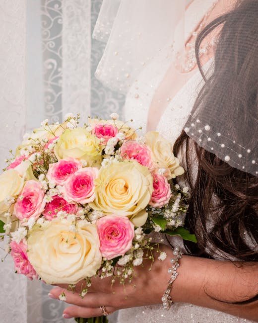 A bride holds a floral bouquet arrangement featuring large cream-colored roses and smaller pink roses, accented with white baby's breath flowers. The bouquet is wrapped in elegant white paper and secu