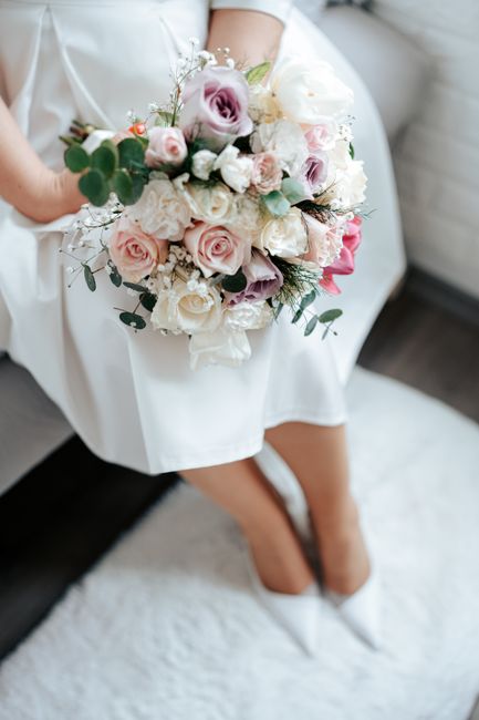 A bride seated on a white upholstered chair, holding a large, lush floral bouquet composed of pale pink and white roses, lavender lisianthus, white peonies, and sprigs of eucalyptus leaves, arranged i