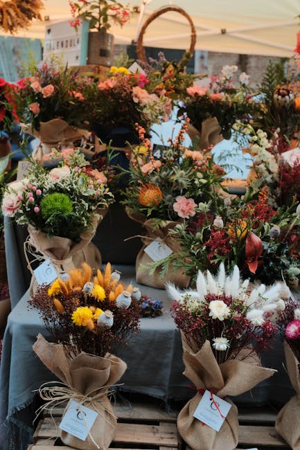 A vibrant display of floral arrangements at a market stall, featuring a variety of fresh flowers including pink roses, white daisies, yellow chrysanthemums, and deep red blooms. The arrangements are w