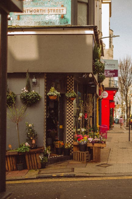 A floral display outside a quaint florist shop on a city street, featuring a bouquet arrangement with a mix of vibrant pink, white, and green flowers placed on a wooden crate, surrounded by potted pla