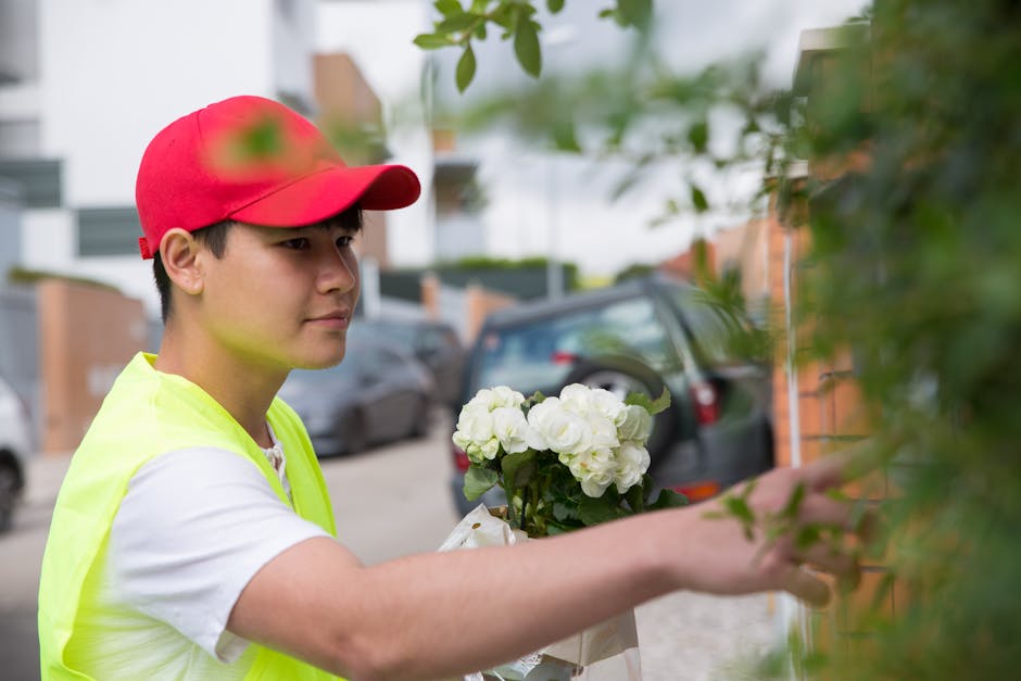 A young delivery person wearing a red cap and high-visibility yellow vest is arranging a bouquet of white hydrangeas and greenery against a brick wall outdoors, demonstrating careful handling of a flo